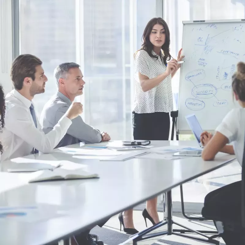 A business woman standing at a white board in a conference room presents to colleagues sitting around a grey modern conference table.