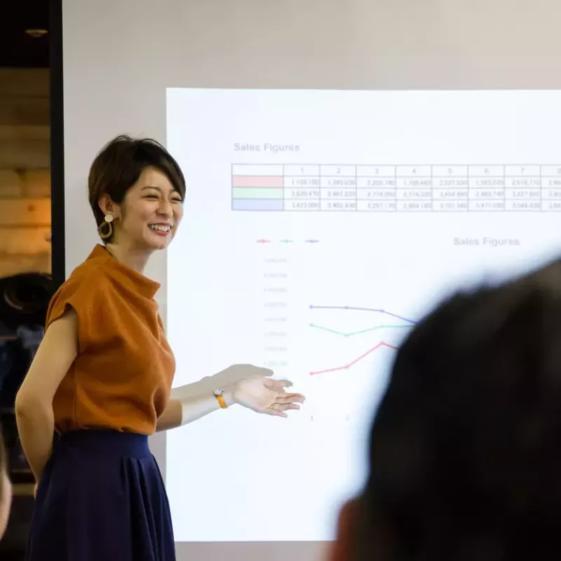 Smiling business woman presents projected sales figures in front of a screen.