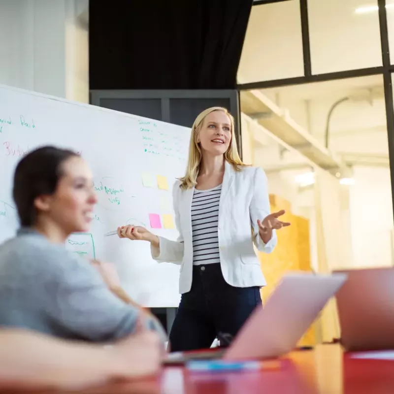 Young business professional in a white blazer speaks at a white board while colleagues smile up at her from a conference table.