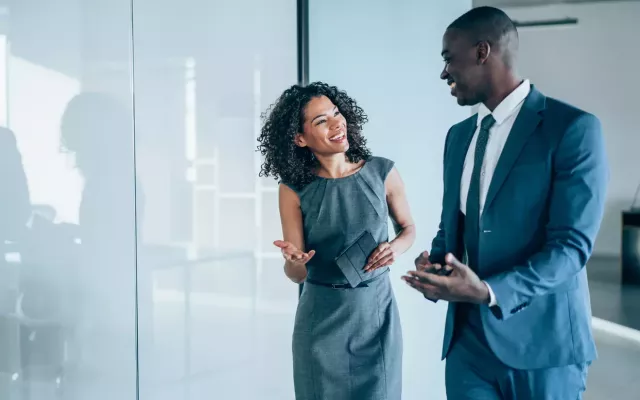 Co-workers in business attire smile and gesture in an office with the glass wall behind them reflecting a conference table and chairs.