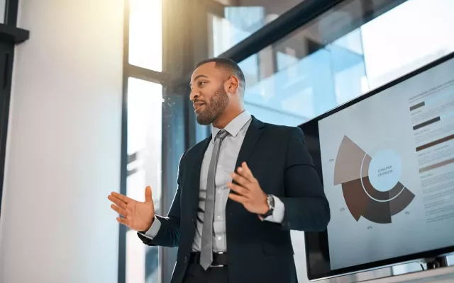 Business professional with a short beard in a suit gestures while presenting in front of a digital screen.