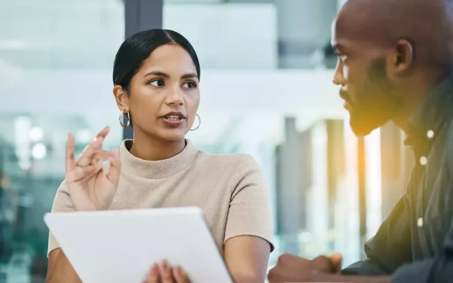 Male and female coworkers discuss sales figures at a conference table in an office with large glass windows with lens flare from sunrise/sunset in background.