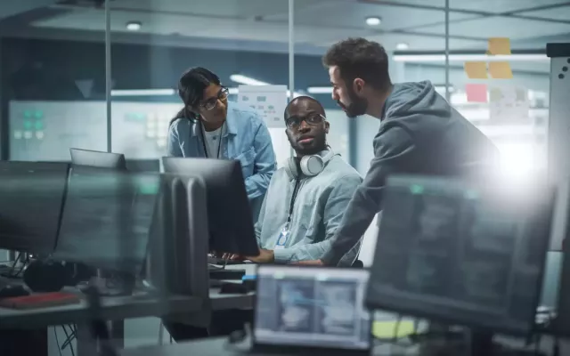 Three coders discuss software improvements in a room full of monitors displaying code.