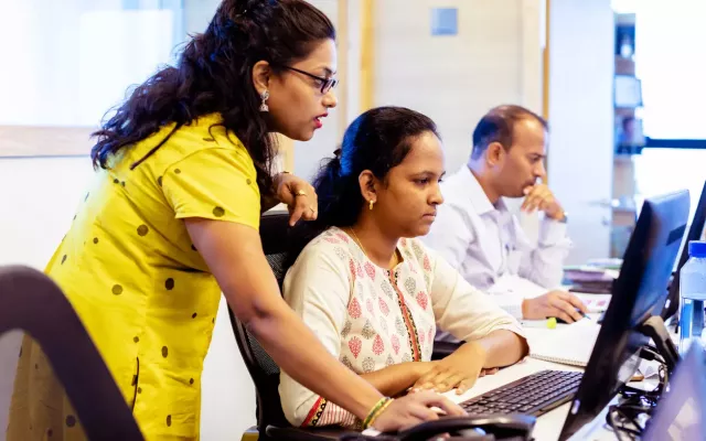 Woman in business casual coaches female colleague at her desk.