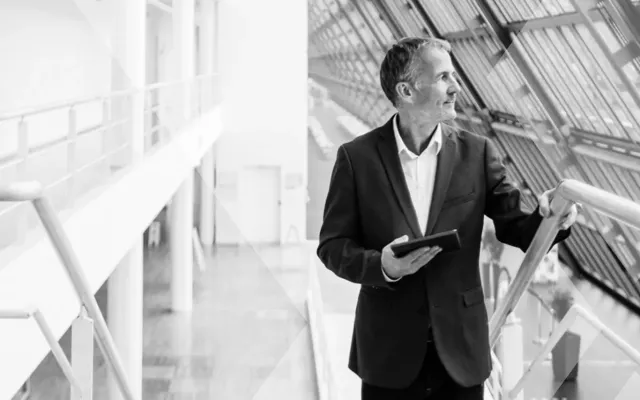 Man in suit jacket look out of large glass windows in a modern office while holding a tablet.