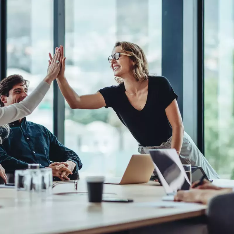 Two women high-five over a conference table with two male colleagues on either side.