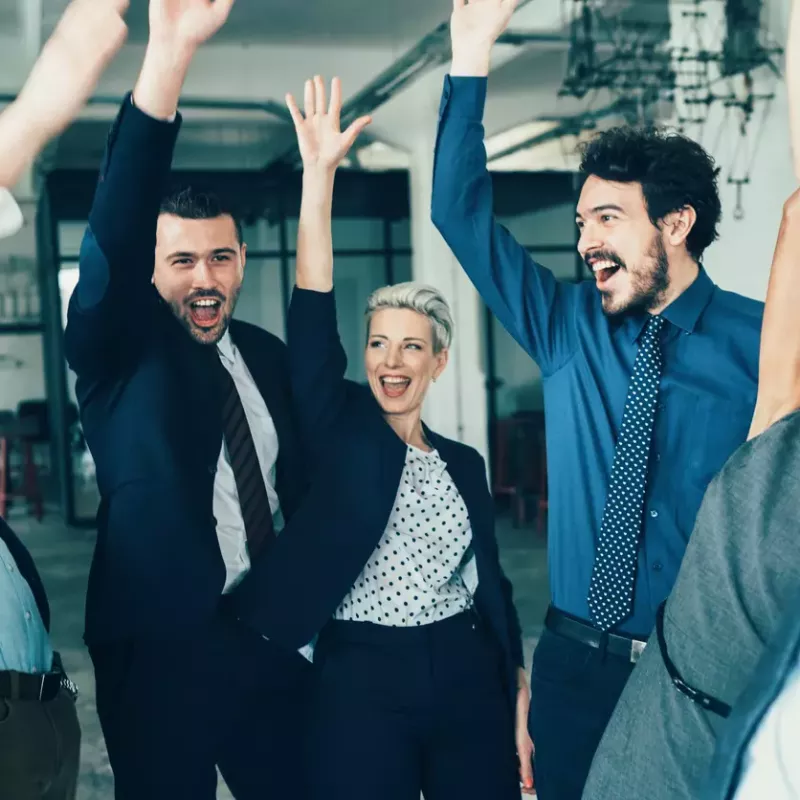 A group of business professionals in a circle celebrates with their hands up.