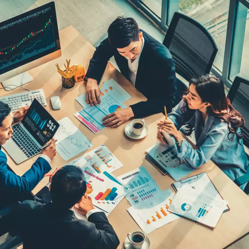 Three male and one female co-worker all in business clothing sit around a wooden table discussing charts and graphs.