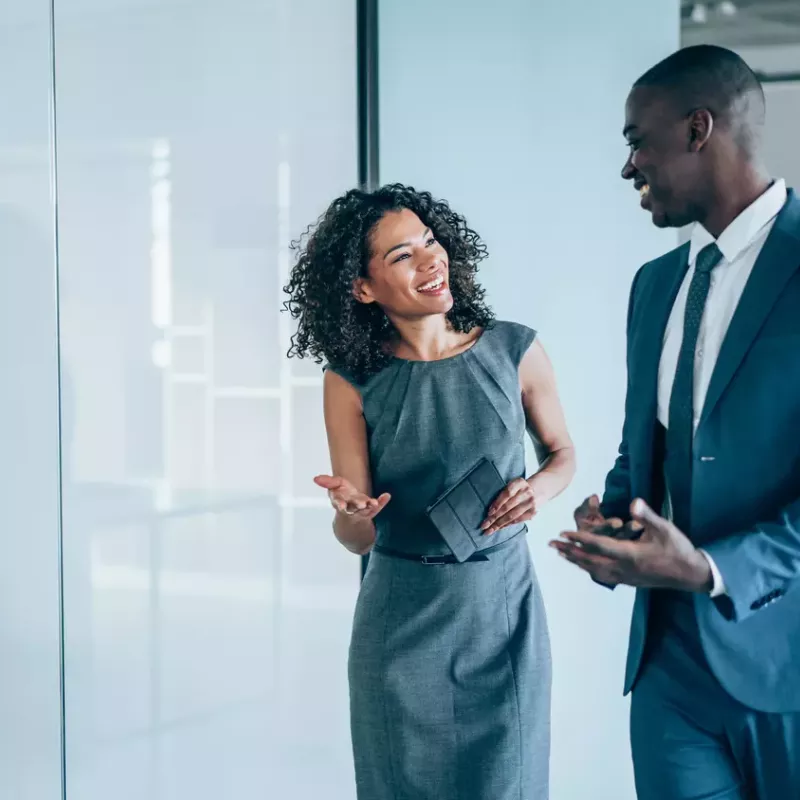 Co-workers in business attire smile and gesture in an office with the glass wall behind them reflecting a conference table and chairs.