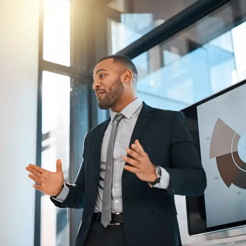 Business professional with a short beard in a suit gestures while presenting in front of a digital screen.