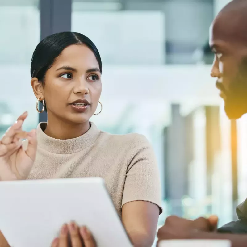 Male and female coworkers discuss sales figures at a conference table in an office with large glass windows with lens flare from sunrise/sunset in background.