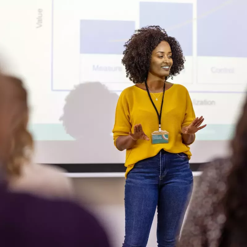 Woman in business casual clothing presents in front of a projector screen showing graphs.