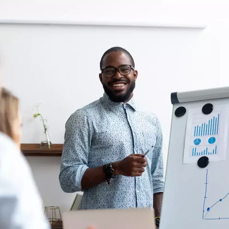 Smiling young professional smiles at his audience while presenting revenue forecasting projections on a whiteboard.