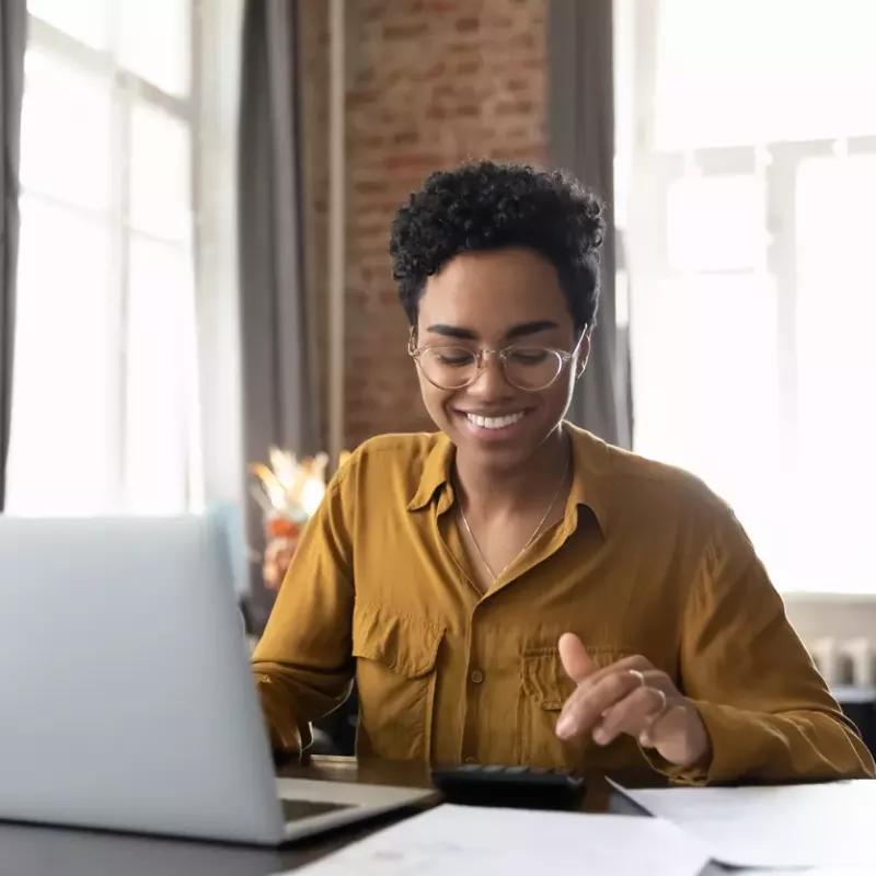 Young black woman wearing glasses and a gold shirt sits at a desk with laptop in a brightly lit room with tall windows and exposed brick walls.