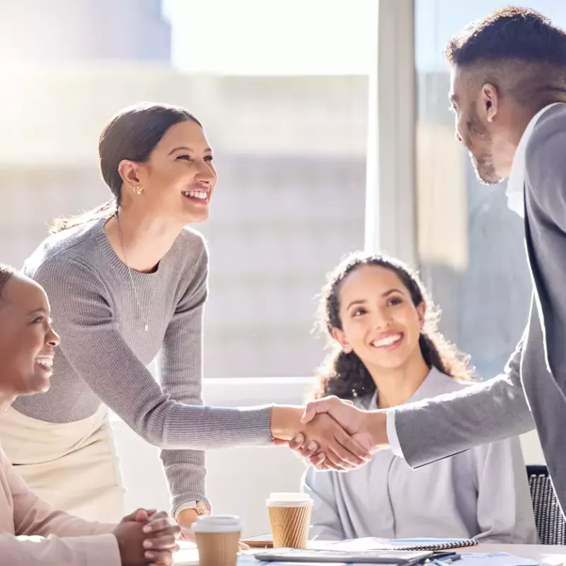 Young professionals shake hands over a conference table in an office with large glass windows.