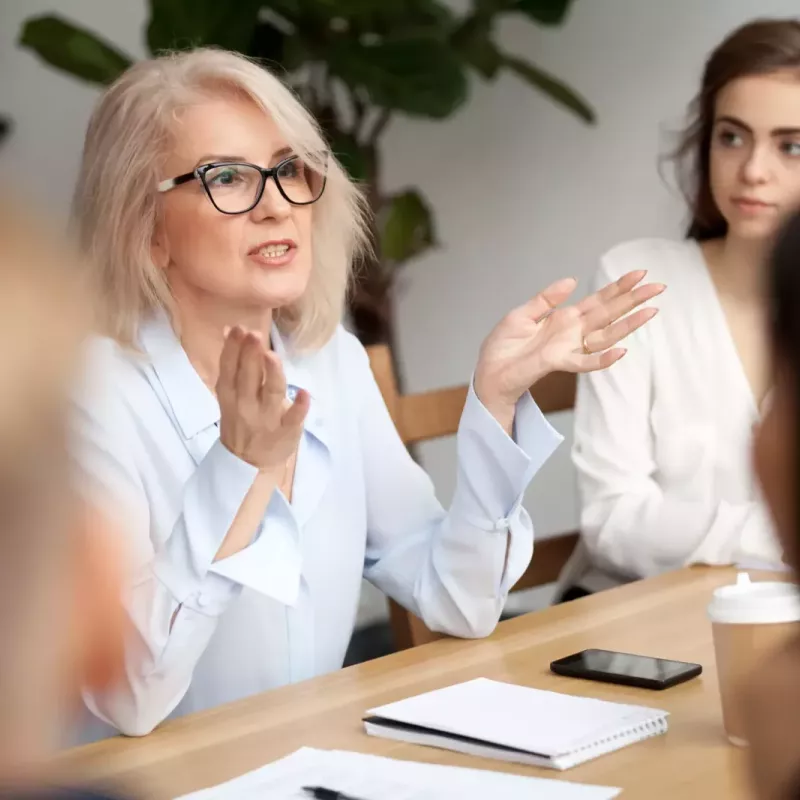 A business leader gestures while sitting at a conference table, speaking to colleagues.