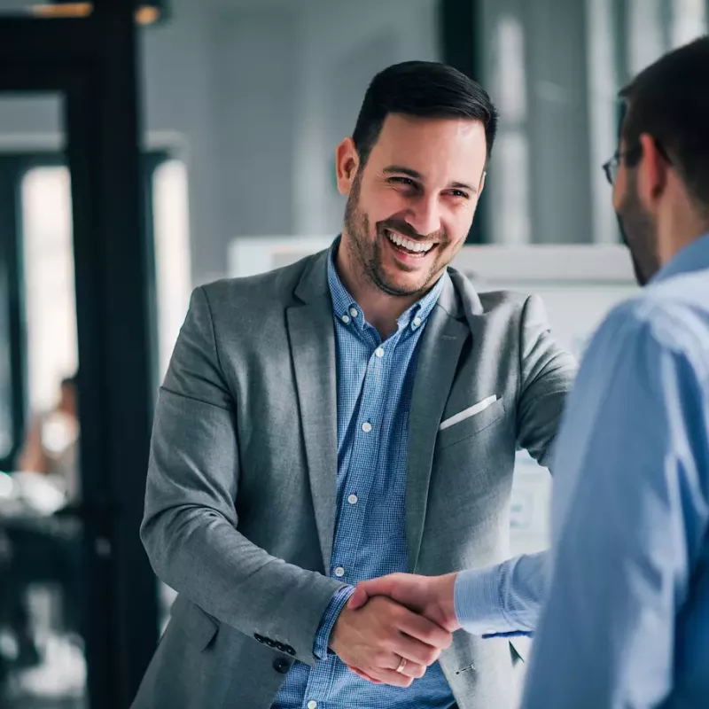 Two young businessmen with short beards shake hands smiling in a modern office.