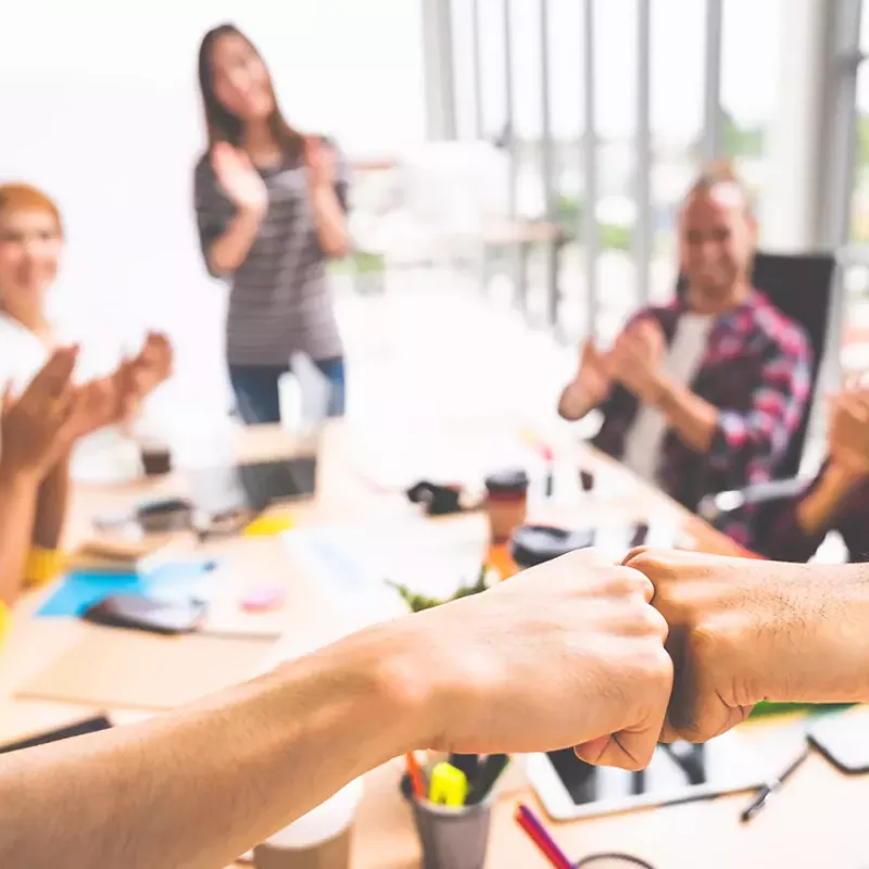 Sales team gathered around a conference table clap in congratulations while two hands fist-bump in the foreground.