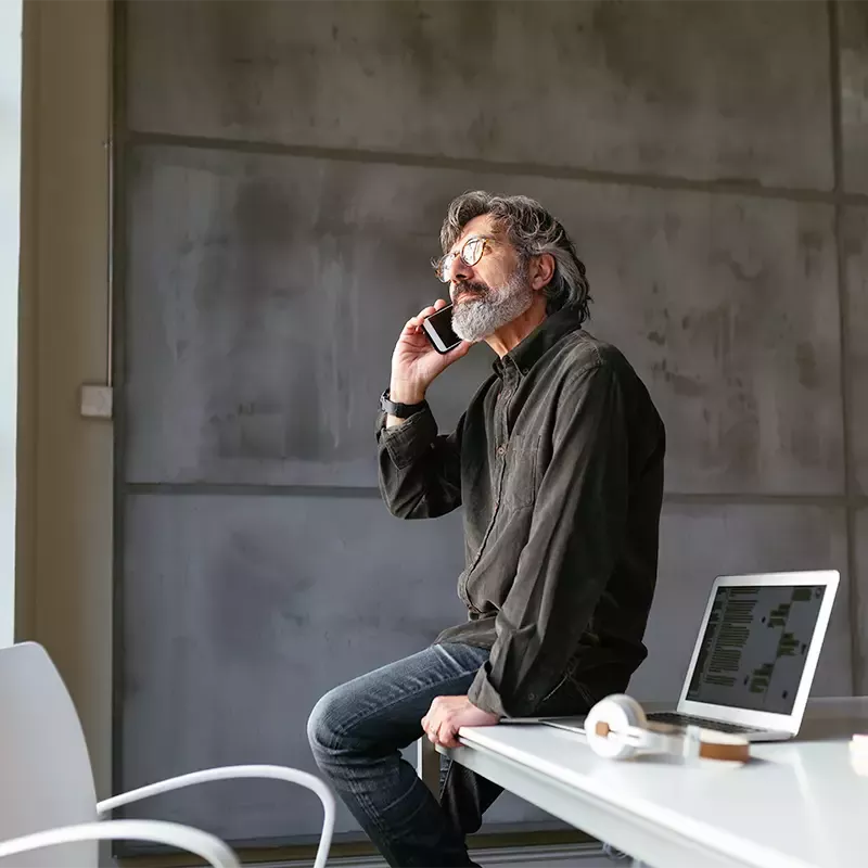 A man in a modern, industrial-style office sits on his desk and looks out the window while talking on the phone.