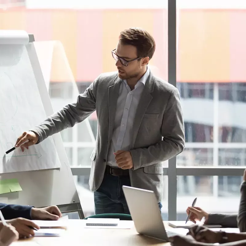 man standing pointing to graph while coworkers around table watch