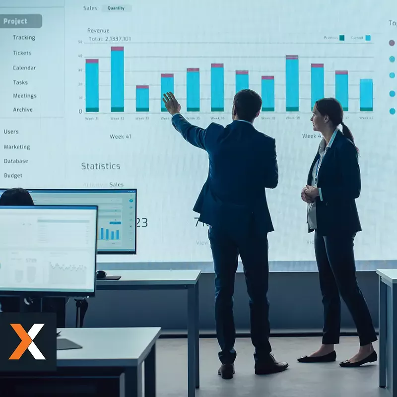 man and woman standing at wall monitor looking at sales performance graphs
