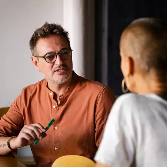 A man in an orange shirt and two other individuals talk at a table in an office or business setting.
