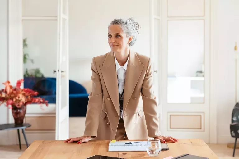 A woman in a tan blazer leans on her arms at the head of a conference table in a small business office with documents in front of her.