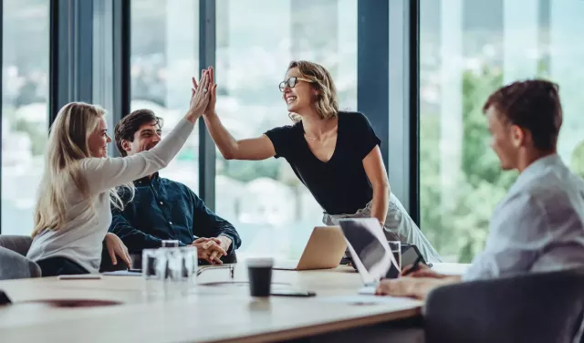 Two women high-five over a conference table with two male colleagues on either side.