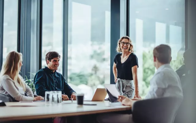 Woman in business casual clothes with glasses leans one hand on a conference table and laughs with coworkers in an office with large glass windows.