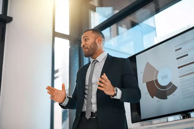 Business professional with a short beard in a suit gestures while presenting in front of a digital screen.