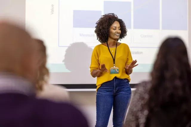 Woman in business casual clothing presents in front of a projector screen showing graphs.