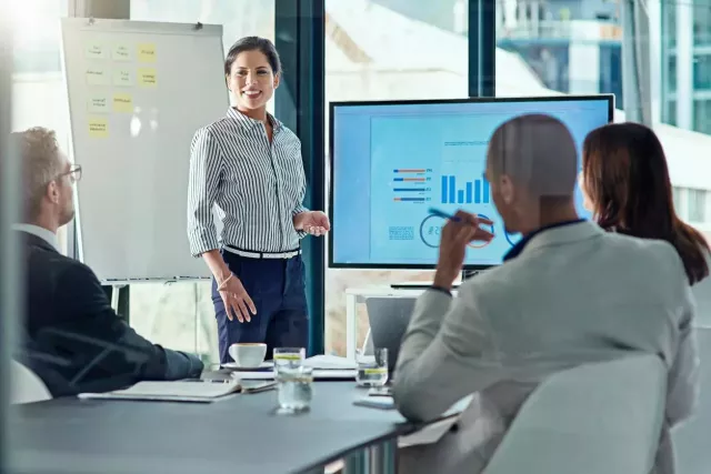 A woman wearing business casual presents sales KPIs on a digital screen in a bright modern conference room.