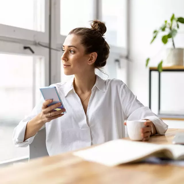 With her cell phone by her side, a woman sits at her desk, fully engaged in her work. She's a modern professional, utilizing technology to stay connected and productive.
