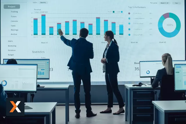 man and woman standing at wall monitor looking at sales performance graphs