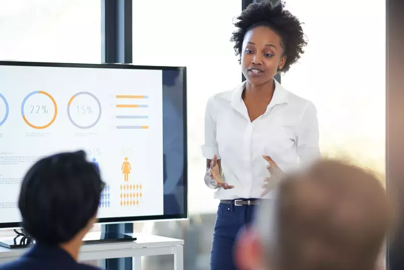 A business woman presents in front of an audience with a screen showing charts and metrics next to her.