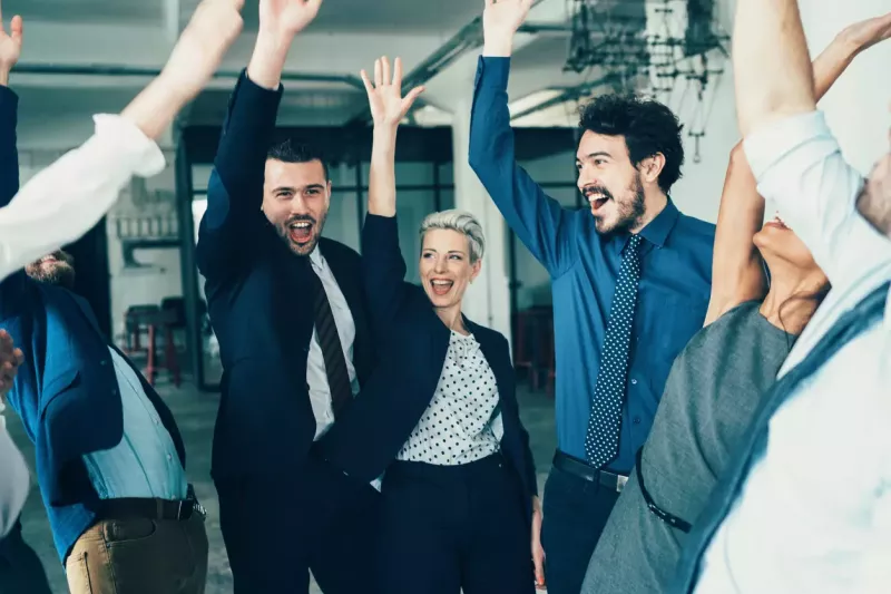 A group of business professionals in a circle celebrates with their hands up.