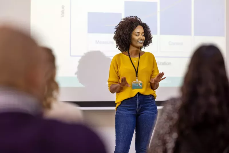 Woman in business casual clothing presents in front of a projector screen showing graphs.