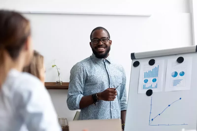 Smiling young professional smiles at his audience while presenting revenue forecasting projections on a whiteboard.