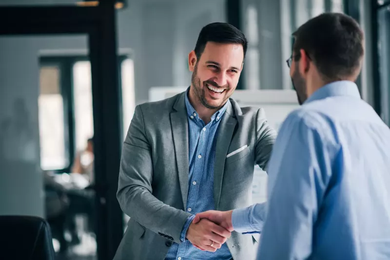 Two young businessmen with short beards shake hands smiling in a modern office.