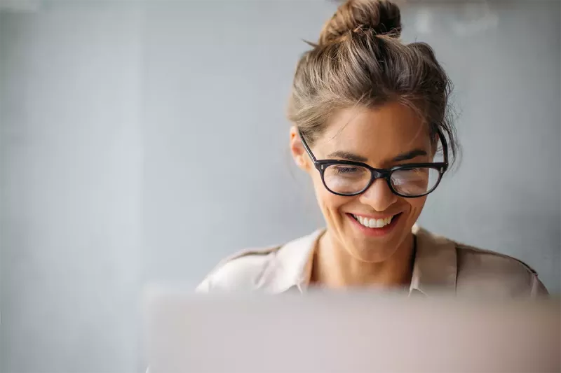 A woman with glasses smiles looking at her laptop computer screen.