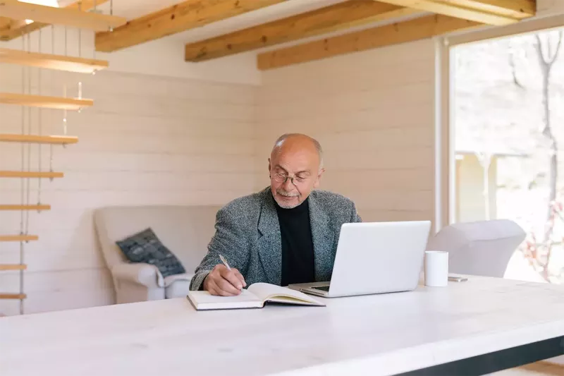 An older gentleman concentrates writing in a notebook while working on a laptop in a rustic environment.