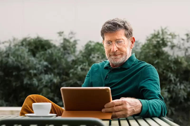 An older man sits outside at a table using a tablet.