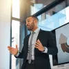 Business professional with a short beard in a suit gestures while presenting in front of a digital screen.