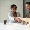 Smiling businesswoman shakes hands with female colleague over a conference table, while male colleagues smiles in the background.