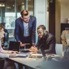 A group of diverse professionals review revenue figures at a table with printed graphs and laptops while smiling.