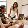 Young colleagues converse with smiles over a conference table in a white office space.