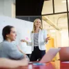 Young business professional in a white blazer speaks at a white board while colleagues smile up at her from a conference table.